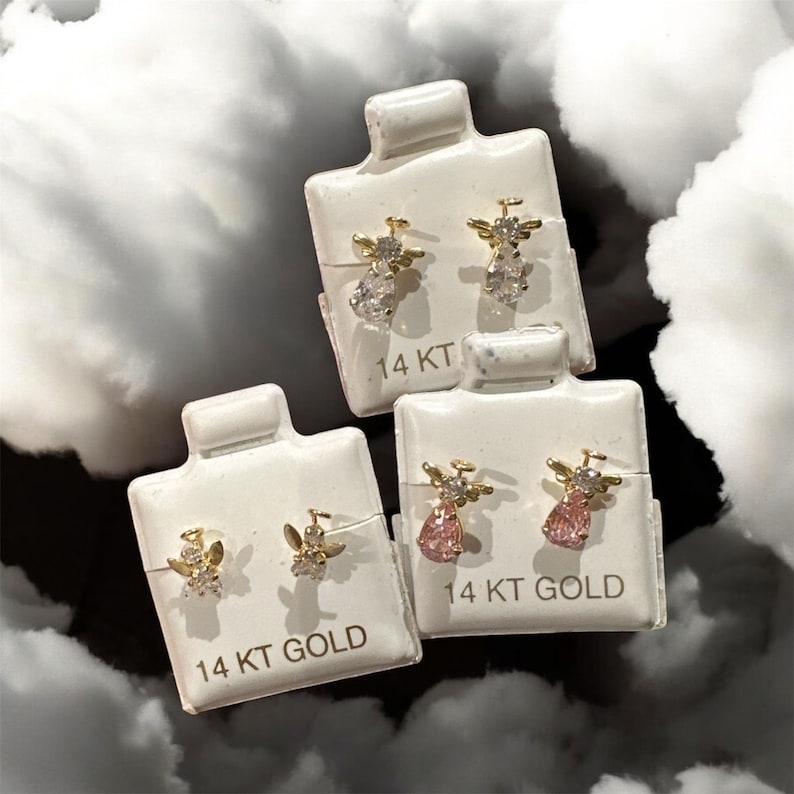 May include: Three pairs of 14 KT gold angel stud earrings on white display cards. Each earring features a gold angel with a clear or pink teardrop-shaped gemstone. The background is a cloudy, dark grey.