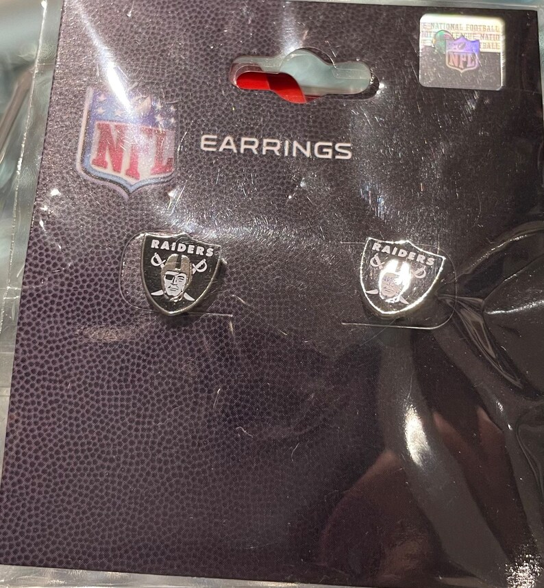 May include: A pair of silver Raiders earrings with the team logo and a player's face on each earring. The earrings are packaged on a black background with the NFL logo and the word "EARRINGS".