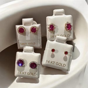 May include: Four pairs of 14KT gold earrings with red gemstones on white display cards. The earrings feature various designs, including round and oval stones, some with surrounding clear stones. The text "14 KT GOLD" is printed on each card.