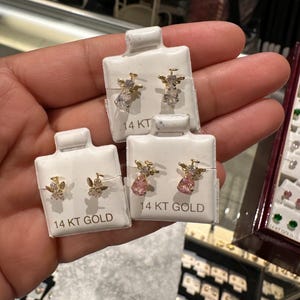May include: Four pairs of 14 KT gold earrings on white display cards. Two pairs feature clear teardrop-shaped gems, while the other two pairs have pink gems. The earrings have a small angel wing design.