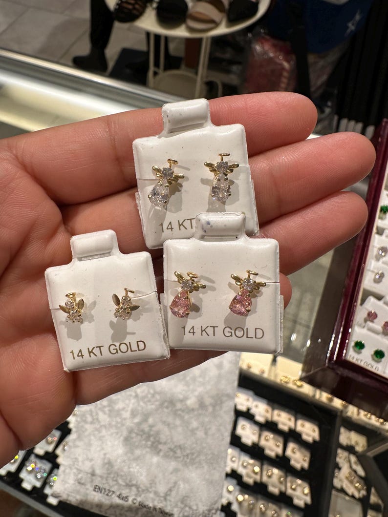 May include: Four pairs of 14 KT gold earrings on white display cards. Two pairs feature clear teardrop-shaped gems, while the other two pairs have pink gems. The earrings have a small angel wing design.