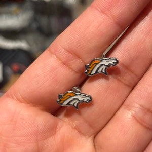 May include: A pair of silver stud earrings featuring the Denver Broncos logo. The logo is white with orange and blue accents.