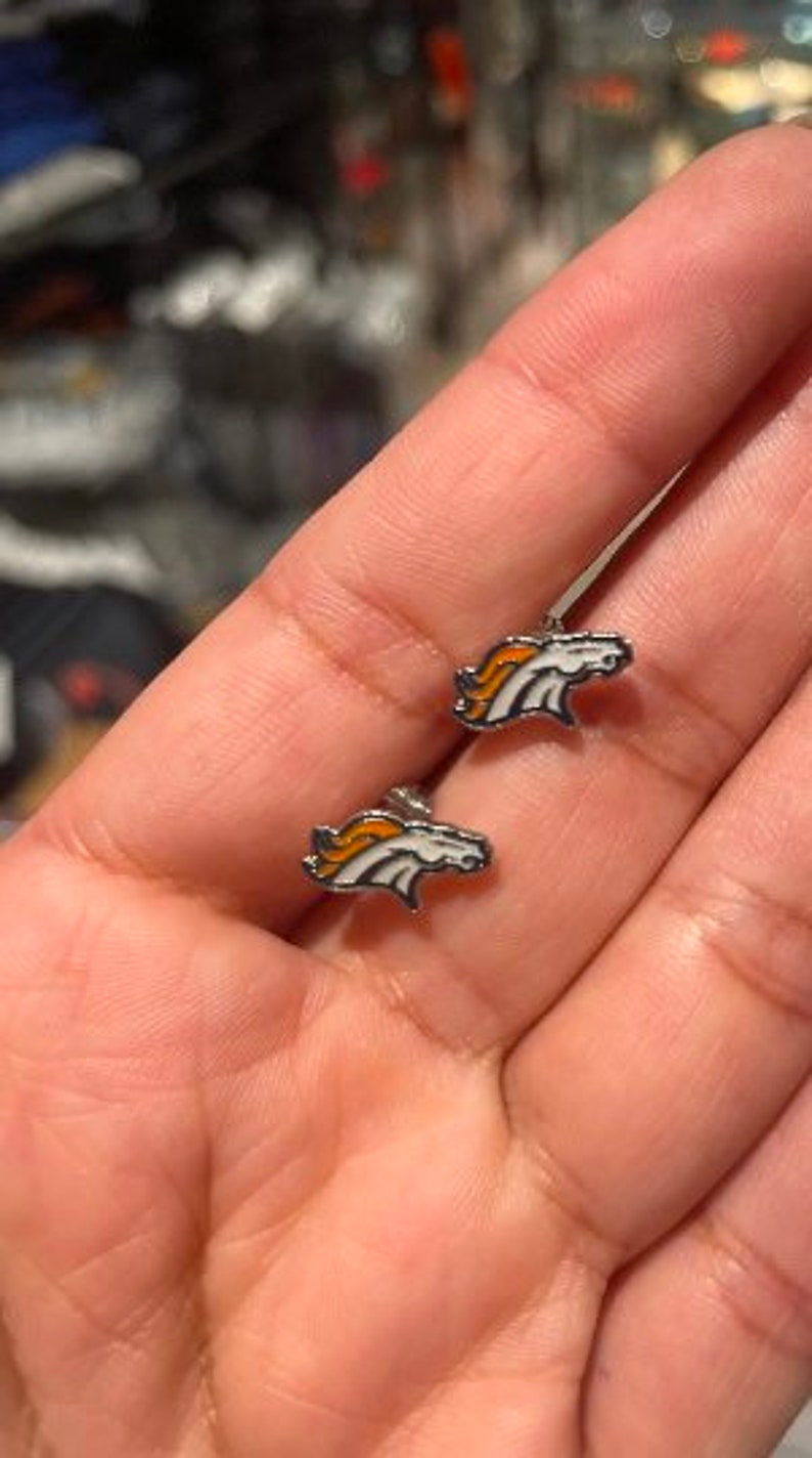 May include: A pair of silver stud earrings featuring the Denver Broncos logo. The logo is white with orange and blue accents.