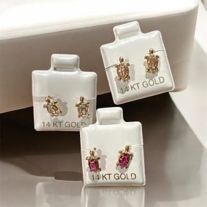 May include: Three pairs of gold-colored turtle stud earrings on white earring cards. Each turtle has a different colored shell: clear, pink, and red. The cards are labeled "14 KT GOLD".