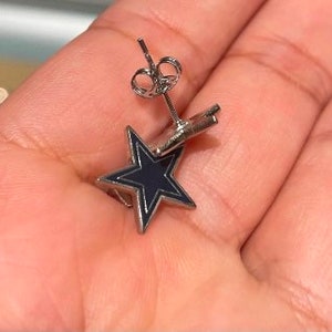 May include: A pair of silver star-shaped earrings with a black enamel fill. The earrings are on a person's hand.