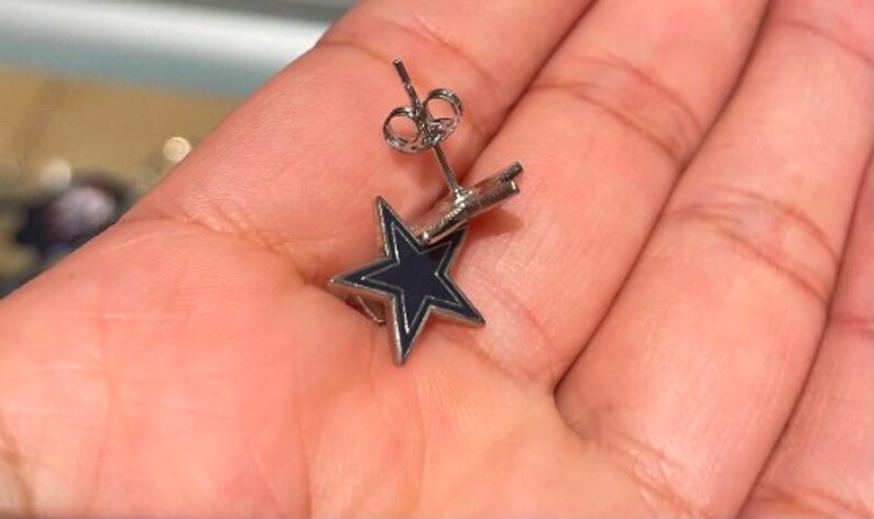 May include: A pair of silver star-shaped earrings with a black enamel fill. The earrings are on a person's hand.