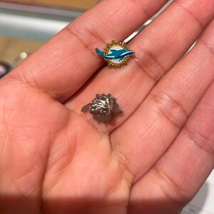 May include: Two small pins, one with a silver dolphin design and the other with a silver and blue dolphin design.
