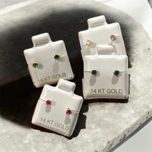May include: Earrings displayed on white cards. The earrings feature various colored gemstones, including green, blue, pink, and gold. The cards are labeled "14 KT GOLD". The earrings are presented on a gray surface.