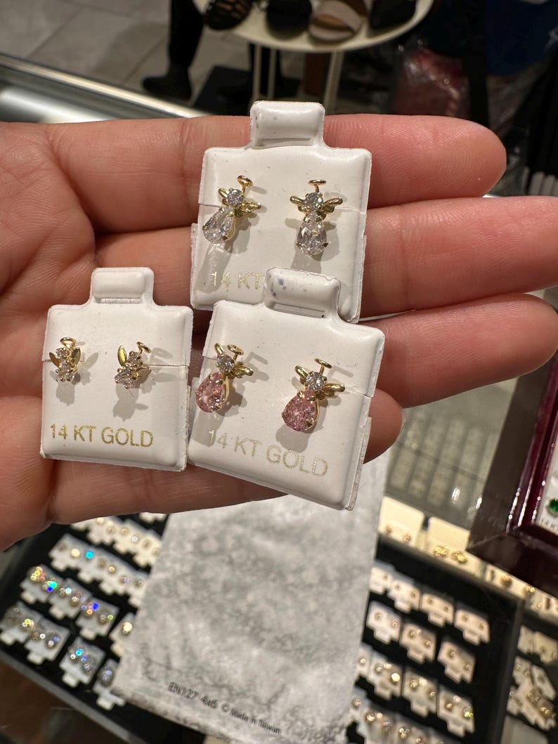May include: Three pairs of 14 KT GOLD angel earrings. One pair features clear teardrop-shaped gems, another has pink pear-shaped gems, and the third has small clear gems. The earrings are displayed on white cards.