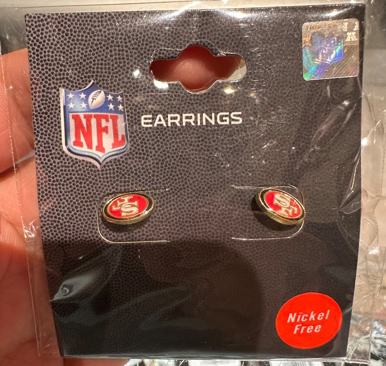 May include: A pair of gold-colored earrings with a red and white San Francisco 49ers logo. The earrings are packaged on a card with the NFL logo and the text "EARRINGS". The card also says "Nickel Free".