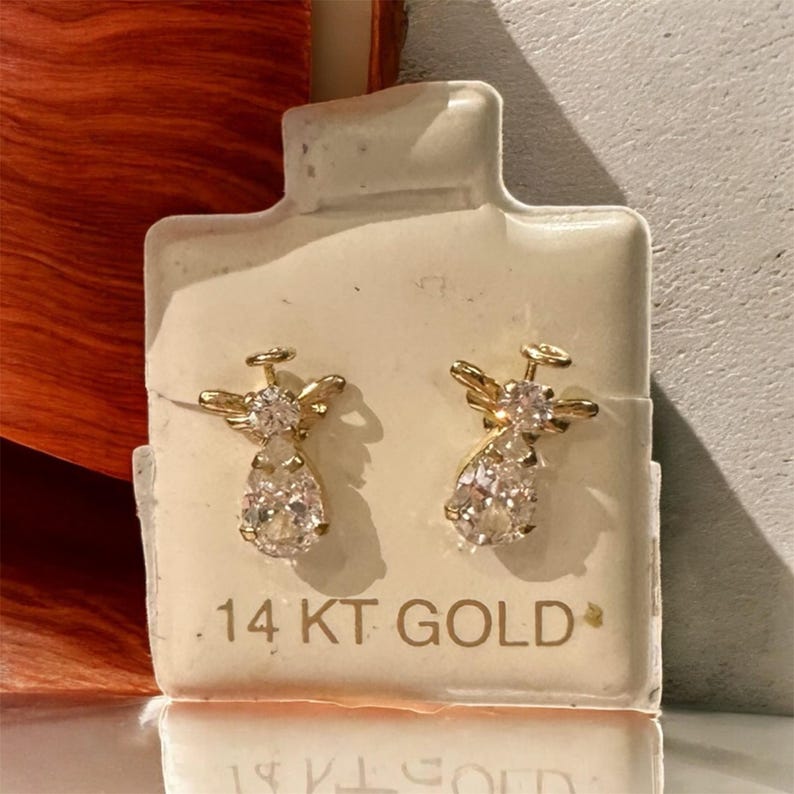 May include: A pair of gold-coloured angel-shaped earrings with clear, teardrop-shaped gemstones. The earrings are displayed on a white card with the text "14 KT GOLD". The earrings have a halo and wing design.