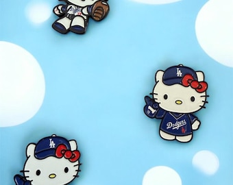 May include: Three Hello Kitty character pins in baseball uniforms. Each pin features Hello Kitty wearing a blue baseball cap with a red bow, and a Dodgers uniform. One pin holds a baseball glove and ball. The background is light blue with white circles.