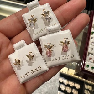 May include: Three pairs of 14 KT gold angel stud earrings on white cards. Each angel has a gold body and wings, with clear or pink gemstone accents. The earrings are displayed in a jewellery store setting.