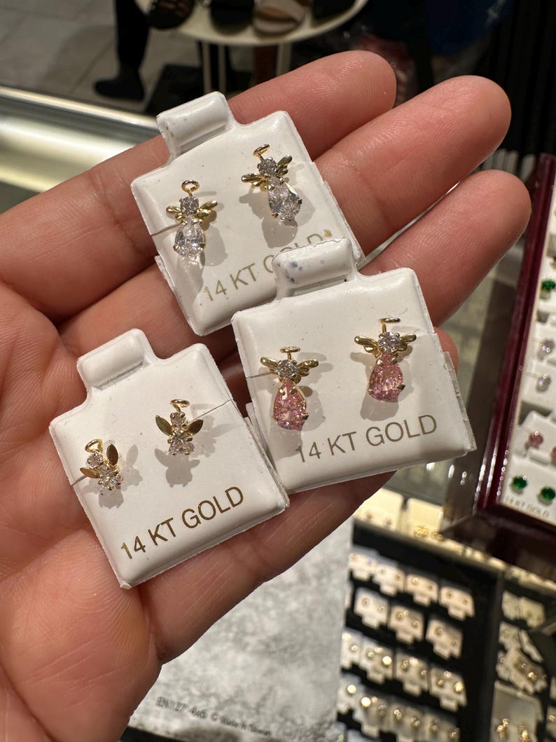 May include: Three pairs of 14 KT gold angel stud earrings on white cards. Each angel has a gold body and wings, with clear or pink gemstone accents. The earrings are displayed in a jewellery store setting.