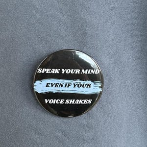 May include: A round, black button with the text "SPEAK YOUR MIND EVEN IF YOUR VOICE SHAKES" in white lettering and a light blue horizontal stripe. The button is pinned to a blue-grey sweatshirt.