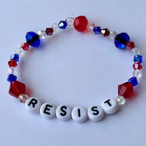 May include: A bracelet made of red, white, and blue beads. The word "RESIST" is spelled out in white letter beads. The beads are of different shapes and sizes, including faceted and round beads. A patriotic accessory.