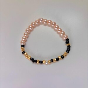 Könnte beinhalten: Ein Perlenarmband mit abwechselnden Abschnitten aus rosa Perlen, goldenen Perlen und schwarzen Perlen. Das Armband wird auf einer weißen Oberfläche präsentiert. Die Perlen sind zu einem flexiblen Band aufgereiht.