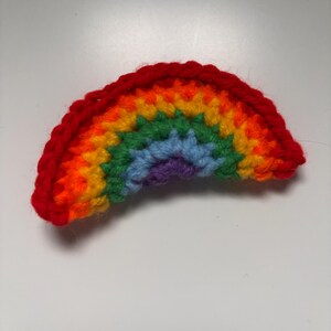 May include: A handmade crochet rainbow in vibrant colors. The rainbow features a red outer band, followed by orange, yellow, green, blue, and purple stripes. The craft item is made with yarn and is set against a white background.