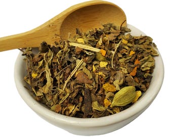 May include: A white ceramic bowl filled with a blend of dried herbs and spices, including green leaves, brown sticks, and yellow turmeric pieces. A wooden spoon is resting on the edge of the bowl.