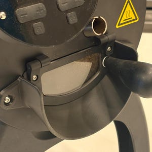 Op de afbeelding: Close-up van een zwarte koffiebrander met een handvat en een kijkvenster. De brander heeft een digitaal bedieningspaneel met knoppen en een waarschuwingssymbool voor hoge temperaturen. De handgreep is zwart en afgerond.