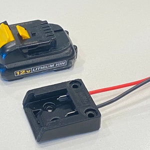 Puede incluir: Una batería de iones de litio de 12 V negra y amarilla con un soporte de batería negro y cables rojo y negro.