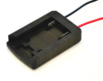 May include: A rectangular black plastic battery holder featuring a recessed centre with two metal contact terminals. Two insulated wires, one black and one red, extend from the side of the component. The housing has a textured surface finish.
