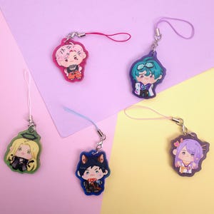 Op de afbeelding: Vijf anime-stijl karakterbedels met lanyards. Elke bedel heeft een ander karakterontwerp met verschillende haarkleuren en outfits. De bedels staan op een pastelpaarse en gele achtergrond.