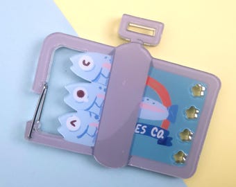 Puede incluir: Accesorio caprichoso en forma de lata de sardinas, de color morado claro. La lata presenta tres peces azules con ojos de dibujos animados y una etiqueta que dice "ES CO". Acentos dorados en forma de estrella adornan la superficie de la lata.