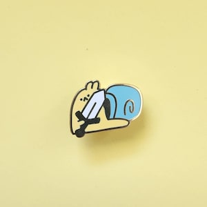 May include: A yellow enamel pin of a cartoon character holding a sword, with a blue swirl behind it.