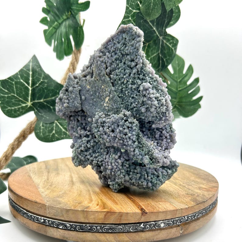 May include: A cluster of grape agate, a unique mineral formation, in shades of lavender and gray. The agate sits on a round wooden display stand with a decorative metal trim. Green leaves and a rope accent the background.