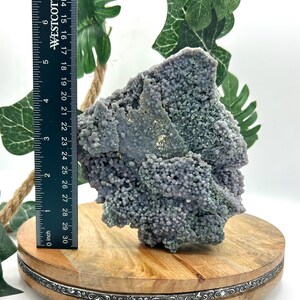 May include: A large, natural grape agate geode specimen. The geode is a mix of lavender and gray colors, with a bubbly, botryoidal texture. A ruler shows the geode is approximately 5 inches tall. The geode sits on a wooden display stand.