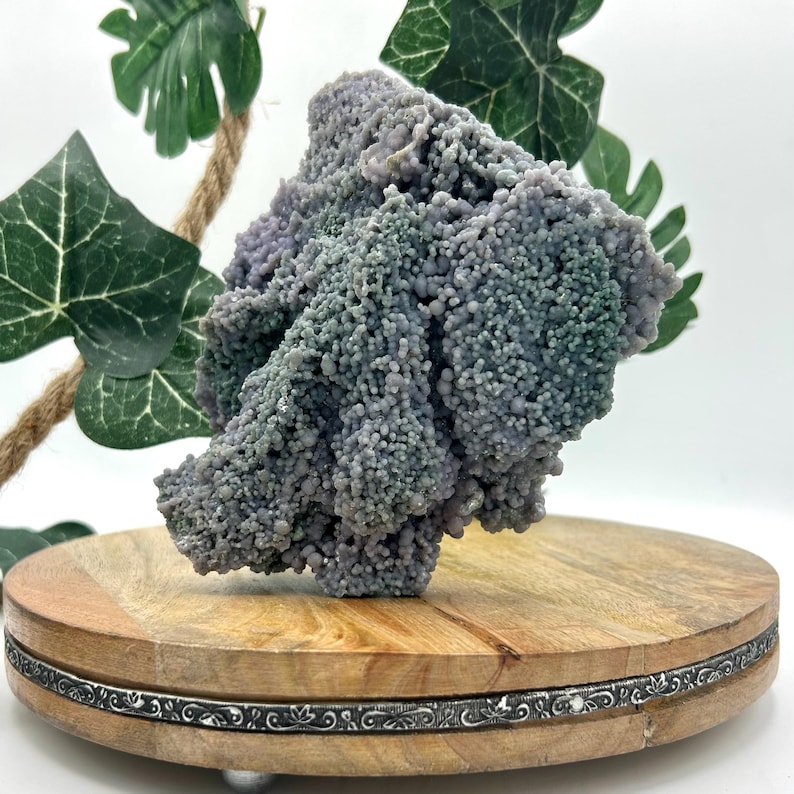 May include: A cluster of botryoidal grape agate, a unique gemstone with a bubbly texture and a mix of lavender and green hues. The agate sits on a round wooden display stand with a decorative silver band.