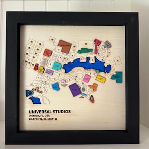 May include: A framed wooden map of Universal Studios in Orlando, Florida, USA. The map features a detailed layout with colorful sections representing different areas and attractions. The text "UNIVERSAL STUDIOS" and coordinates are visible.