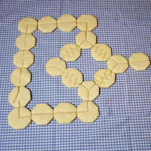 May include: A collection of 18 yellow sugar cookies arranged in a spiral pattern on a blue and white checkered tablecloth. The cookies are decorated with a variety of designs, including geometric shapes and letters.