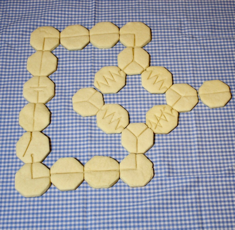 May include: A collection of 18 yellow sugar cookies arranged in a spiral pattern on a blue and white checkered tablecloth. The cookies are decorated with a variety of designs, including geometric shapes and letters.