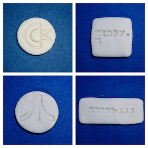 Puede incluir: Cuatro cortadores de galletas de cerámica blanca con diferentes diseños. El primer cortador es un círculo con la letra "C" dentro. El segundo cortador es un rectángulo con la palabra "READY" dentro. El tercer cortador es un círculo con un diseño de invasor espacial. El cuarto cortador es un rectángulo con las palabras "ERROR 110" dentro.