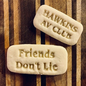 May include: Two white rectangular biscuits with text. The first biscuit says "Hawkins AV Club" and the second biscuit says "Friends Don't Lie".