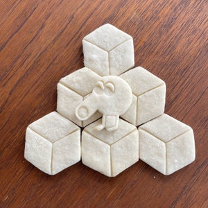 May include: A set of six white cookie cutters shaped like cubes and a cartoon animal. The cutters are arranged in a triangular pattern with the animal in the centre.