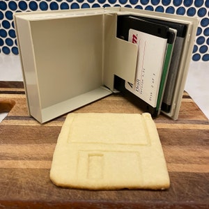 May include: A white plastic floppy disk case with three floppy disks inside. The case is open and the disks are visible. The disks are labelled "Draft Version 3.21 Disk 2 of 3". A cookie shaped like a floppy disk is on a wooden surface.