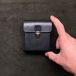 Op de afbeelding: Een zwart leren etui met een zilveren sluiting. Het etui is gesloten en heeft een glad, gestructureerd oppervlak.