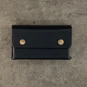 May include: A black leather wallet with two brass snaps. The wallet has a fold-over flap and a smooth, textured surface.