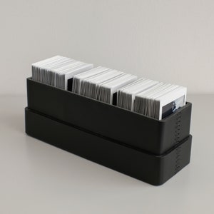 Op de afbeelding: Een zwarte opbergdoos met drie stapels witte fotokaarten erin. De doos heeft een gestikte rand.