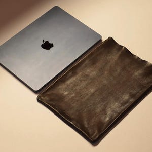 May include: A closed, silver laptop with a black Apple logo next to a dark brown leather laptop sleeve. The sleeve has a textured surface and a slightly wavy edge. The items are on a light beige surface.