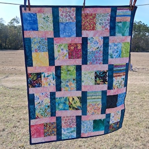 May include: A colorful patchwork quilt with a variety of floral and abstract patterns. The quilt features squares of fabric in shades of blue, pink, green, and yellow, connected by teal blue strips. The quilt is hanging on a clothesline outdoors.