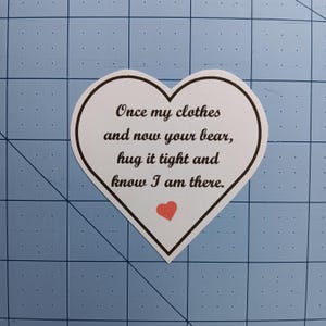 May include: White heart-shaped card with a black border and a red heart in the center. The card has the text "Once my clothes and now your bear, hug it tight and know I am there."