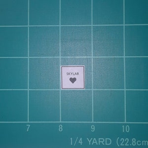 May include: A small, square white label with the name "SKYLAR" printed in black capital letters above a black heart symbol. The label is centered on a green cutting mat with grid lines and measurement markings.