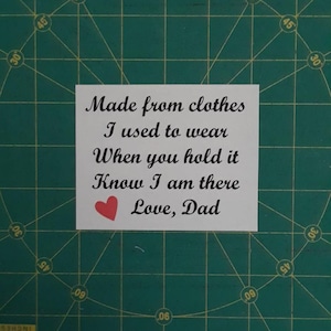 May include: A white rectangular label with black text that reads "Made from clothes I used to wear. When you hold it Know I am there. Love, Dad." A red heart is below the text. The label is on a green surface.