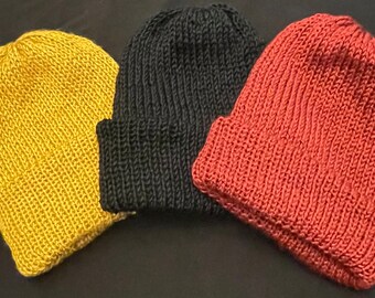 Puede incluir: Tres gorros de punto en diferentes colores: amarillo mostaza, negro y naranja óxido. Los gorros están doblados y dispuestos sobre una superficie oscura.