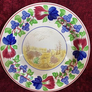 May include: A white ceramic plate with a blue and green floral border. The center of the plate features a hand-painted scene of rabbits in a field.