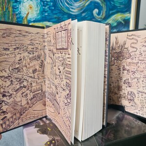 May include: An open book with detailed map illustrations on the inside covers. The pages are white, and the book is on a dark surface. The map is in brown and beige tones, featuring intricate drawings.